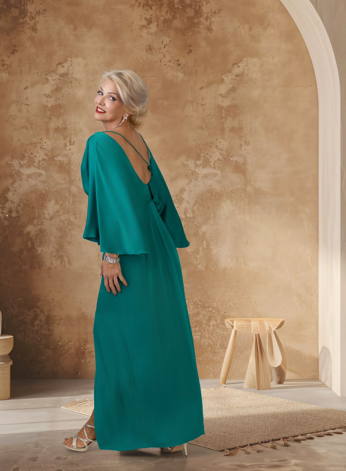 Woman in flowing emerald green dress with cape sleeves poses elegantly against textured beige wall with arched doorway.