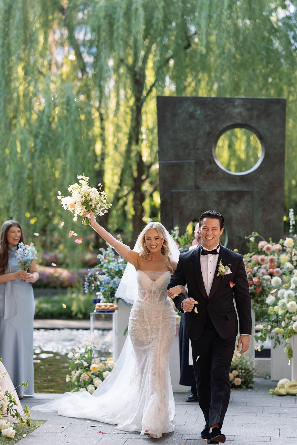 A joyful wedding ceremony exit under weeping willow trees with floral arrangements and a modern circular arch backdrop.