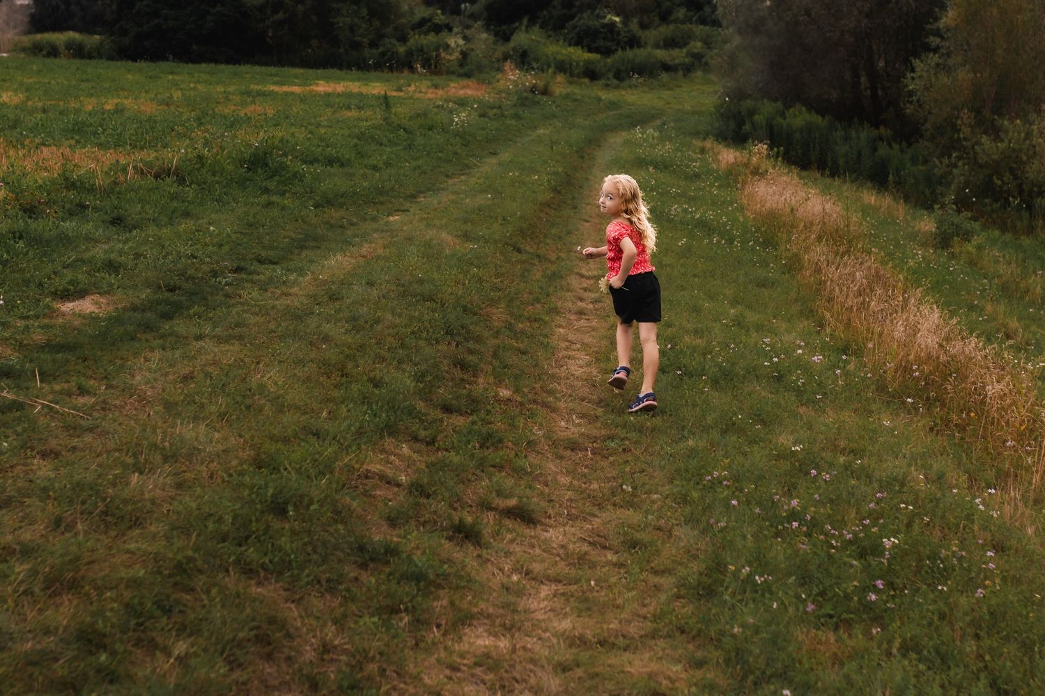 Runner in pink top and black shorts follows a grassy trail path through a green countryside field on a sunny day.