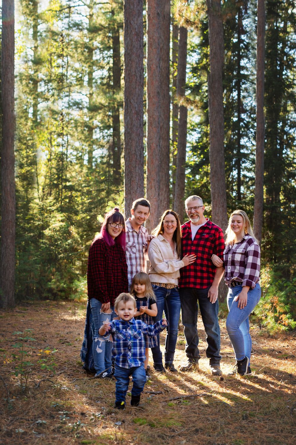 A group wearing matching plaid flannel shirts poses together in a sunlit forest of tall pine trees.