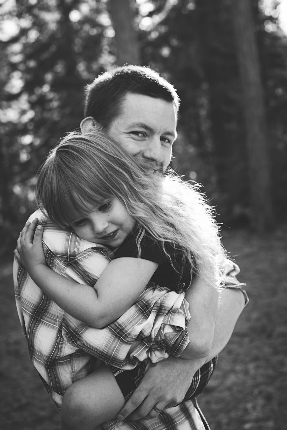 A tender black and white portrait of a father and child embracing outdoors with trees in the background.