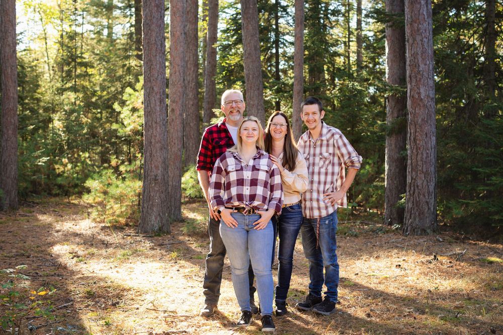 Family wearing coordinated red plaid shirts poses together in a sunlit forest among tall pine trees.
