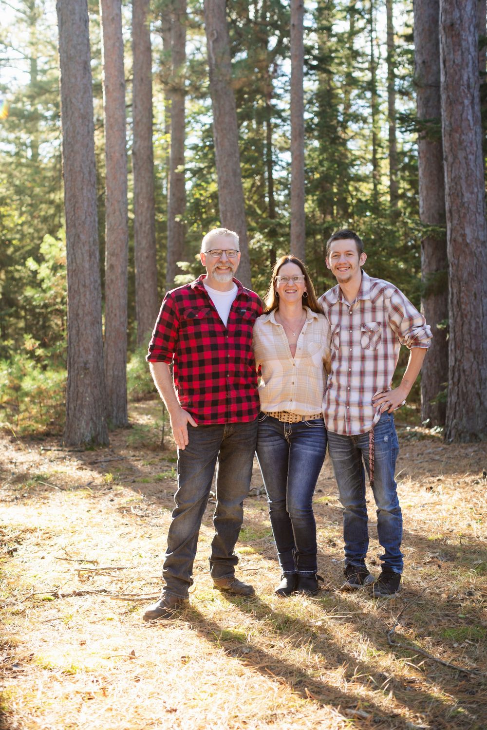 A group stands casually in a sunlit forest with tall pine trees, wearing plaid shirts and denim.