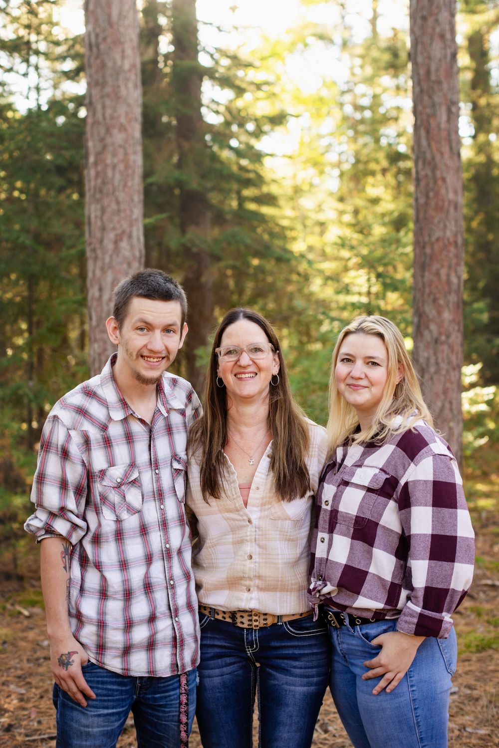Three people in plaid shirts and jeans pose together in a forest setting during golden hour.