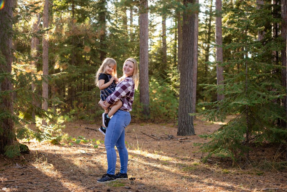 A person holding a child stands in a sunlit forest path surrounded by tall evergreen trees.
