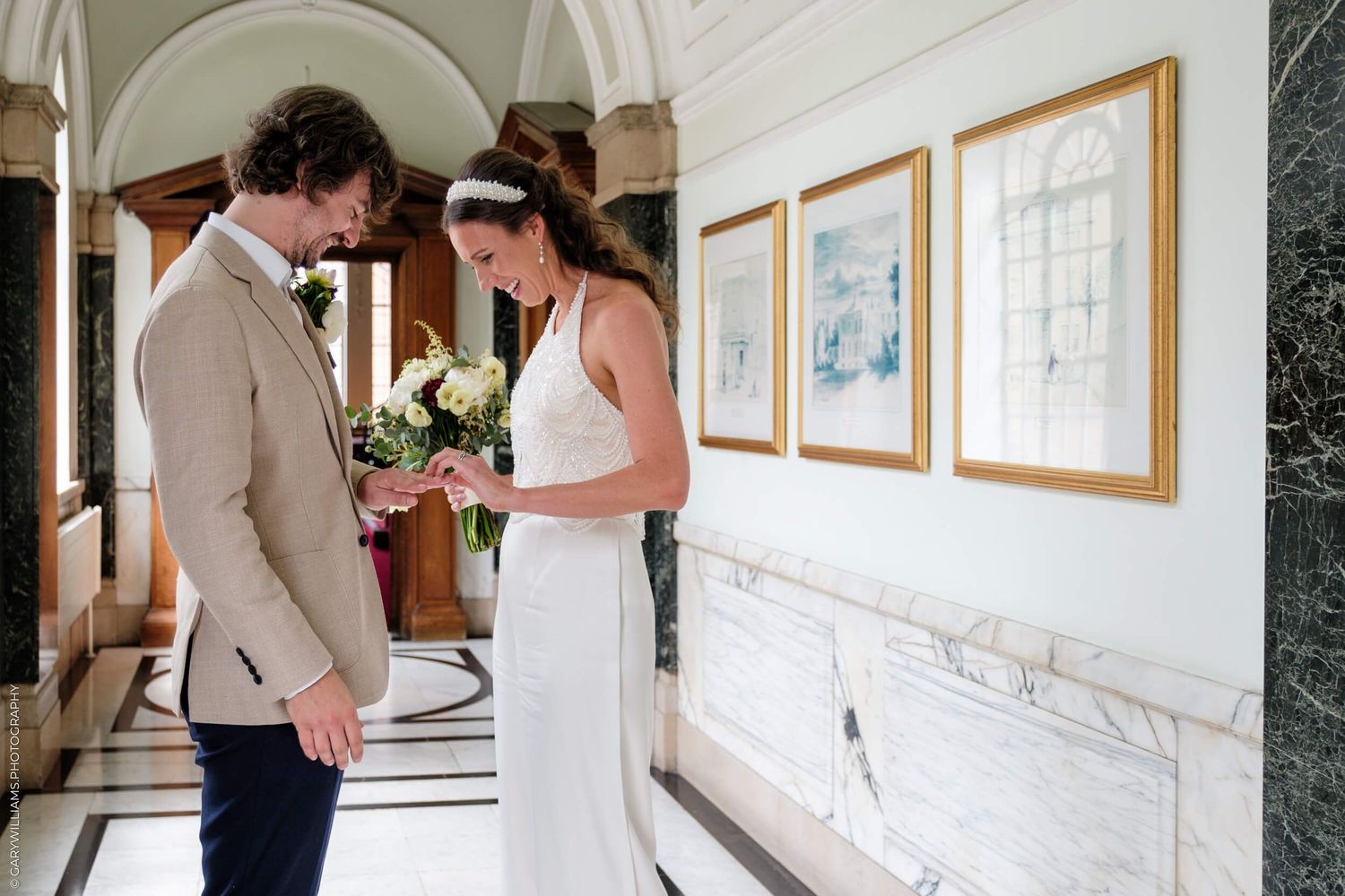 A couple in elegant wedding attire shares a quiet moment in a hallway with framed artwork on the white walls.