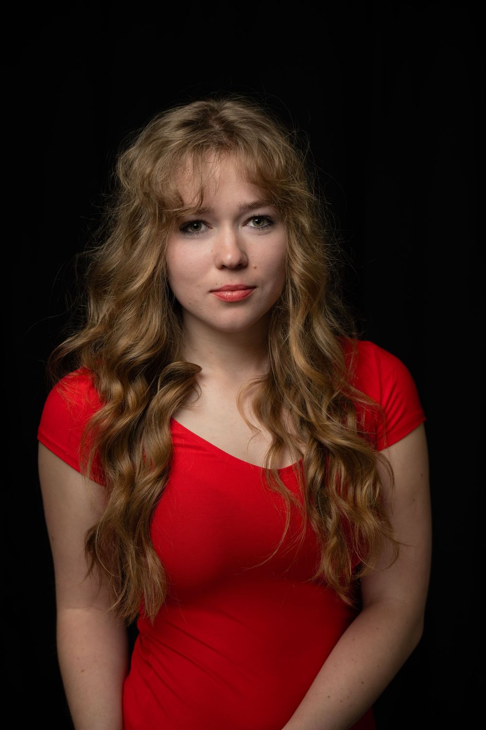 An actress in a vibrant red dress poses for a headshot in a professional photography studio against a black backdrop.