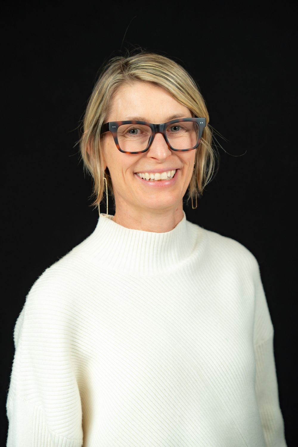 Professional headshot of someone wearing glasses and a white sweater smiling against a black background.