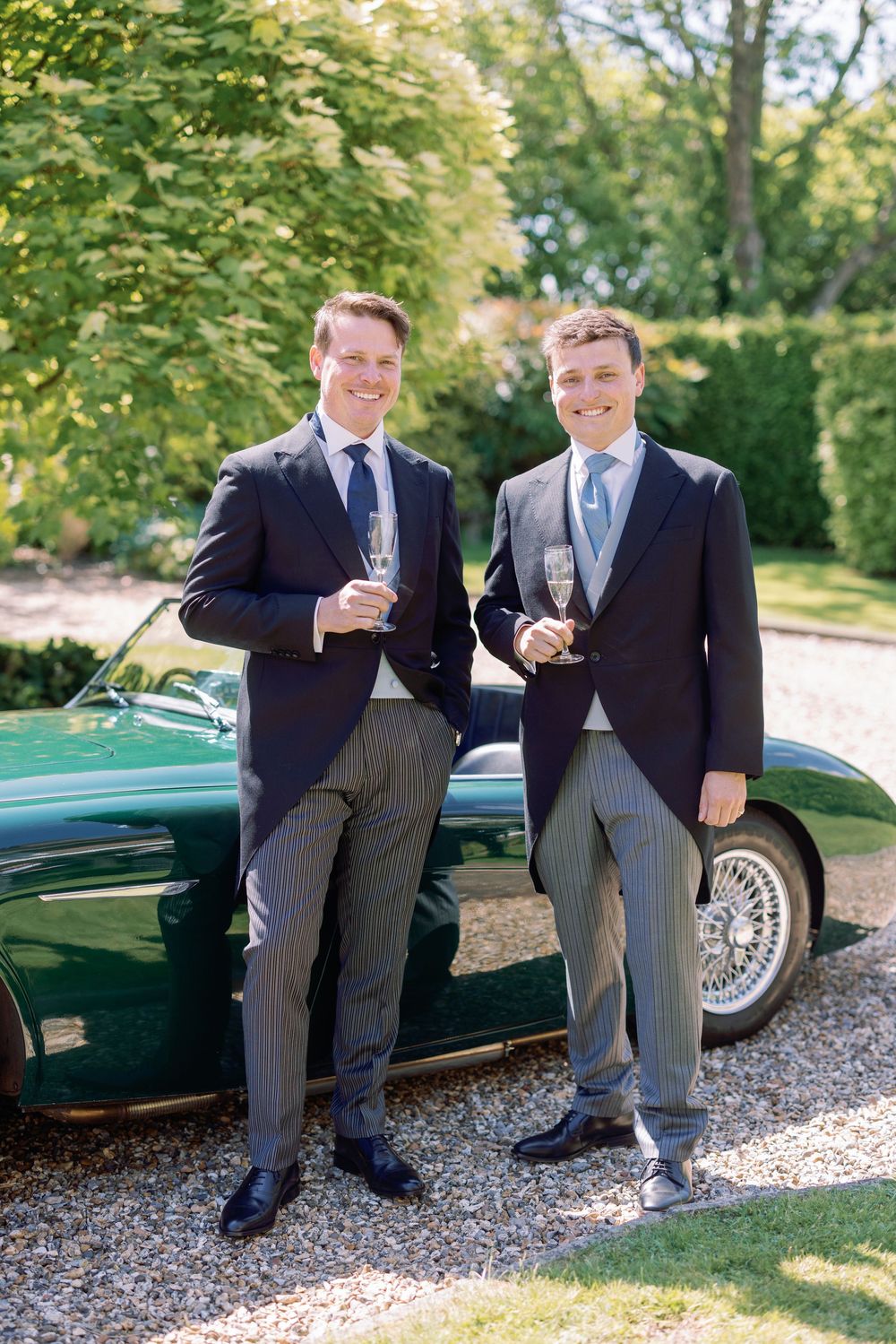 Two people in formal attire raise wine glasses while leaning against a classic British racing green sports car.