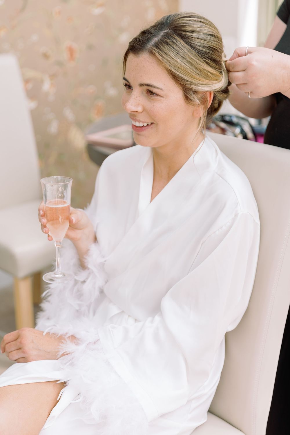 A person wearing a white robe sits smiling while holding a glass of rose wine during hair styling preparations.