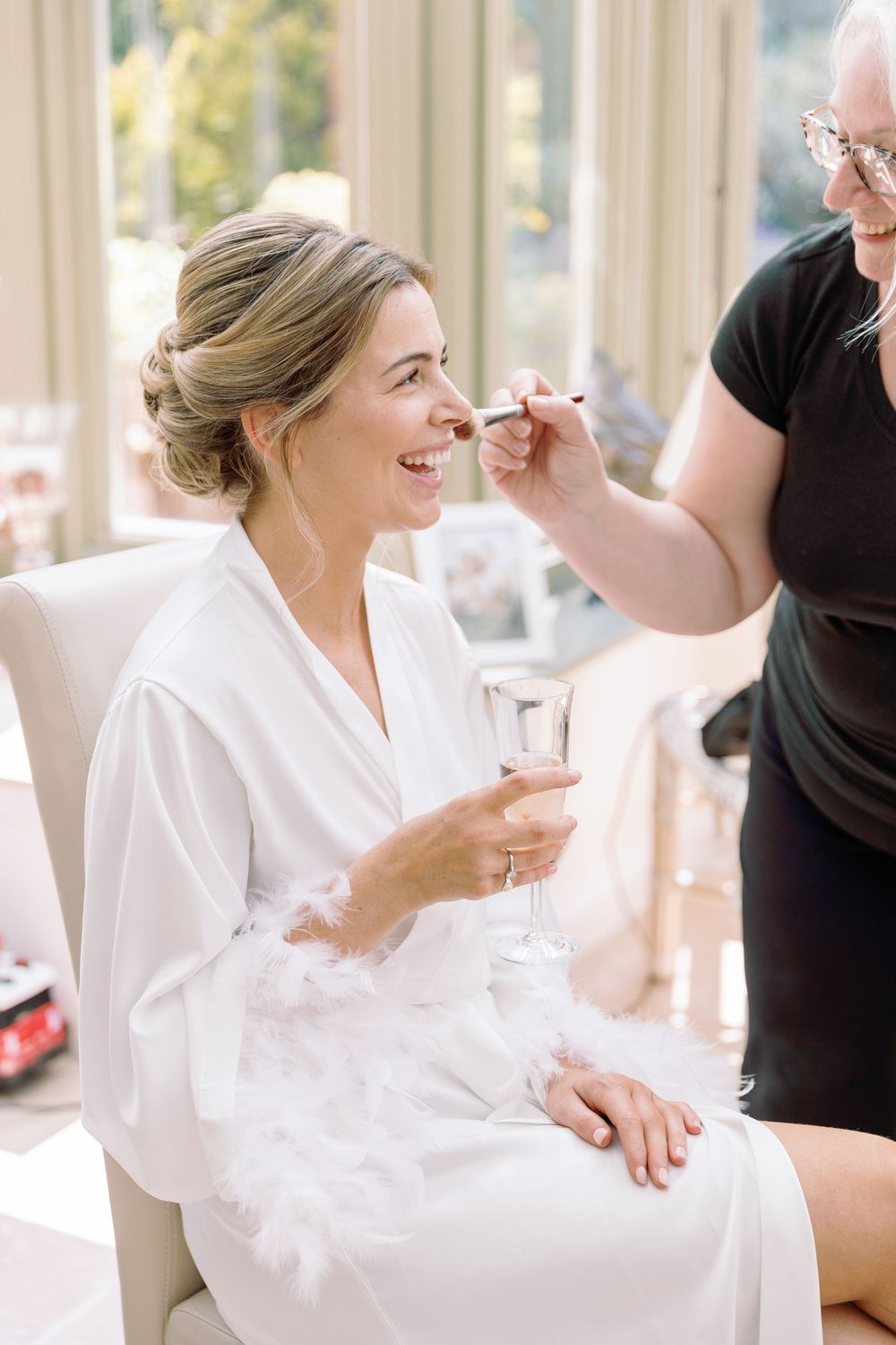 Person in white robe having makeup applied while sitting in bright sunlit room.