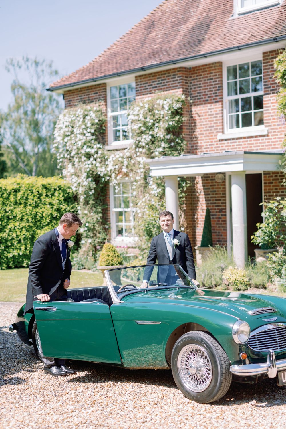 Two people admiring vintage green Austin Healey convertible parked outside brick country house.