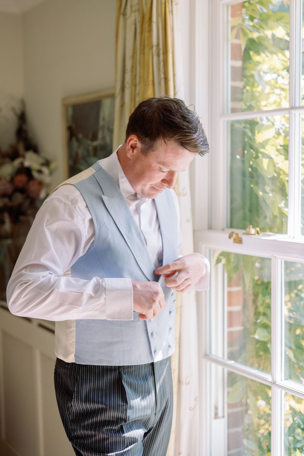 Groom in light blue vest and striped pants adjusts his outfit by a sunlit window.