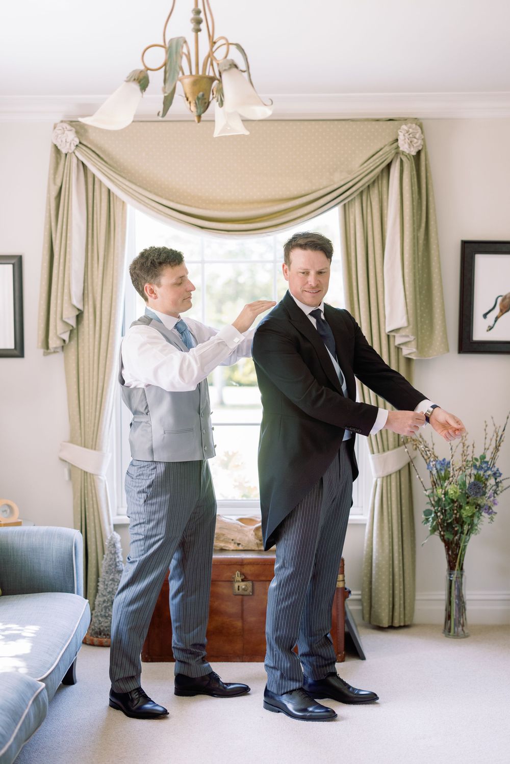Two people getting ready in an elegant room with sage green curtains and a chandelier.