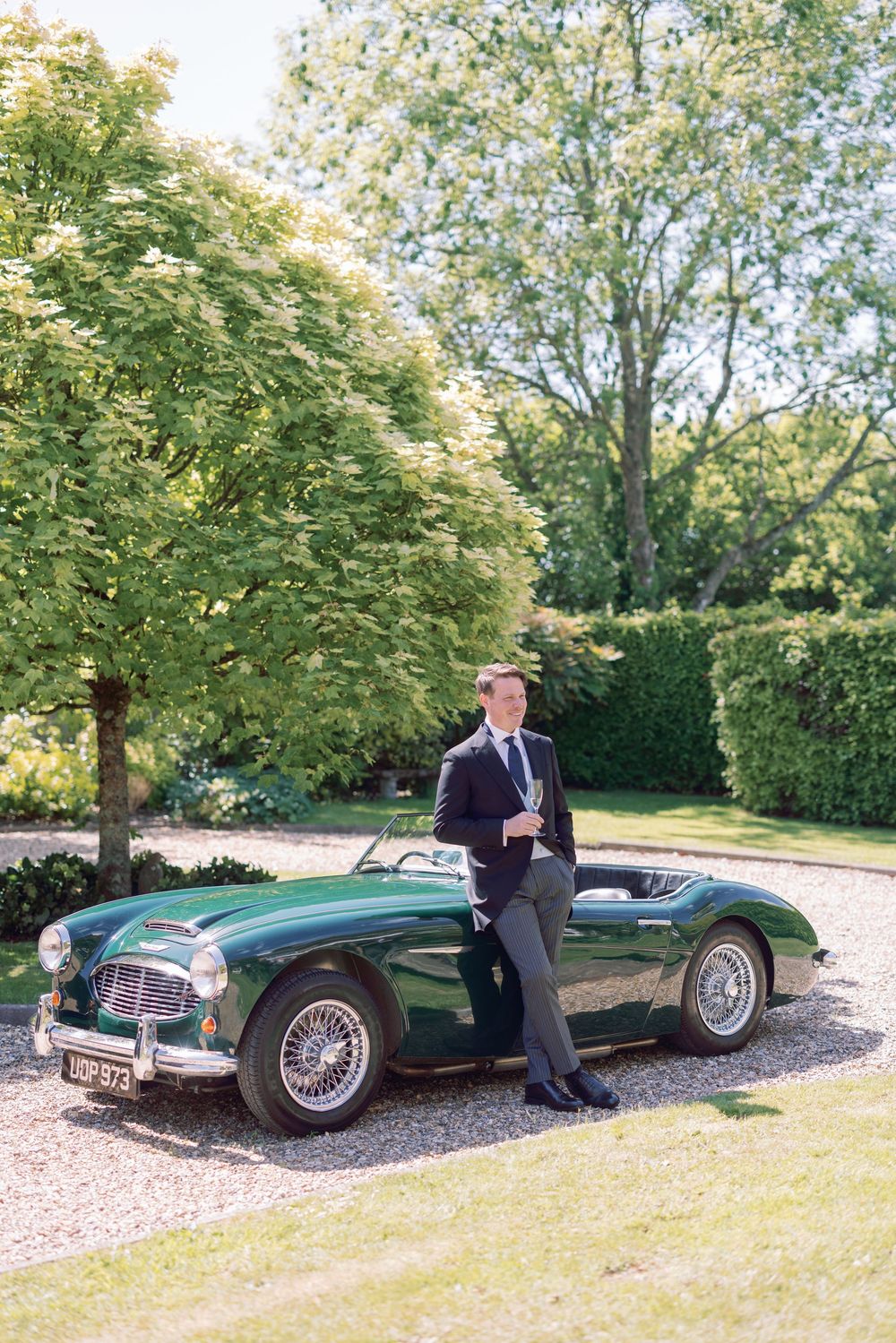 Classic green Austin Healey sports car parked on gravel driveway beside garden.