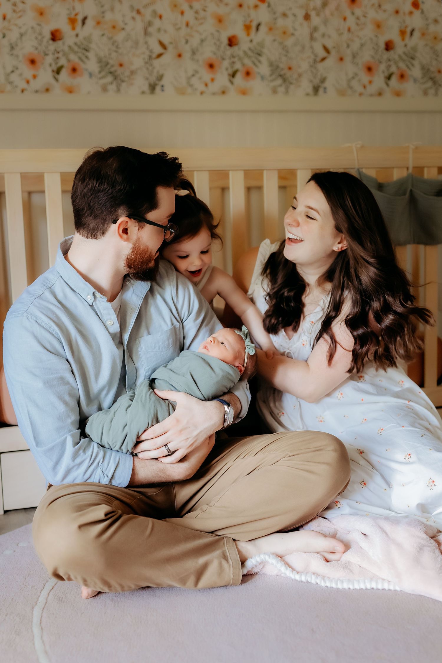 A joyful family moment shared on a bed with a newborn baby in a light blue outfit against a floral wallpaper background.