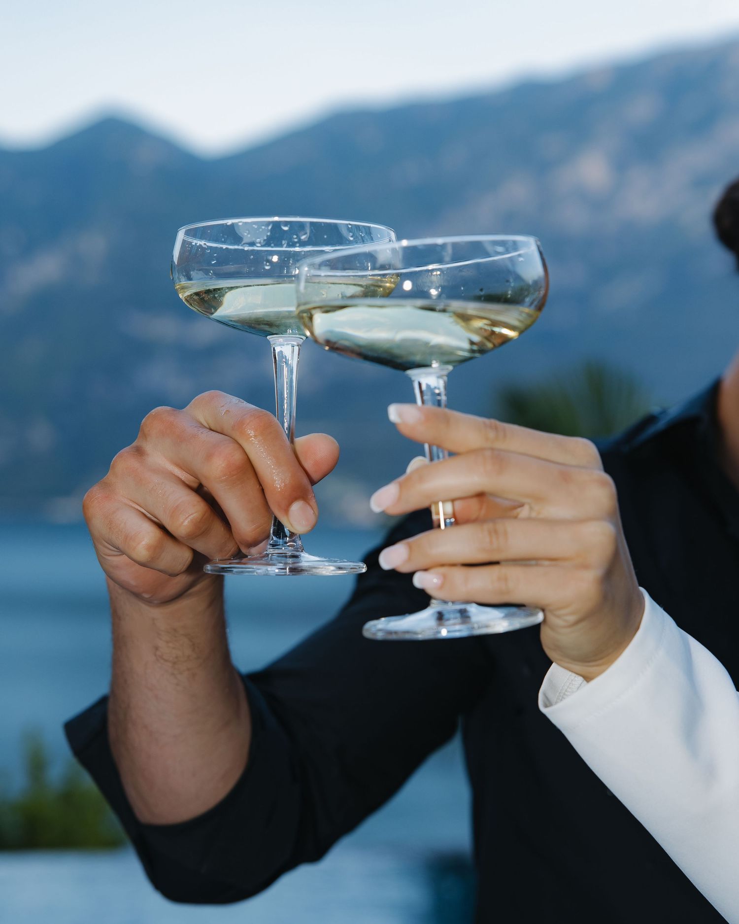 Two hands toasting with champagne coupe glasses against a backdrop of mountains and a lake.