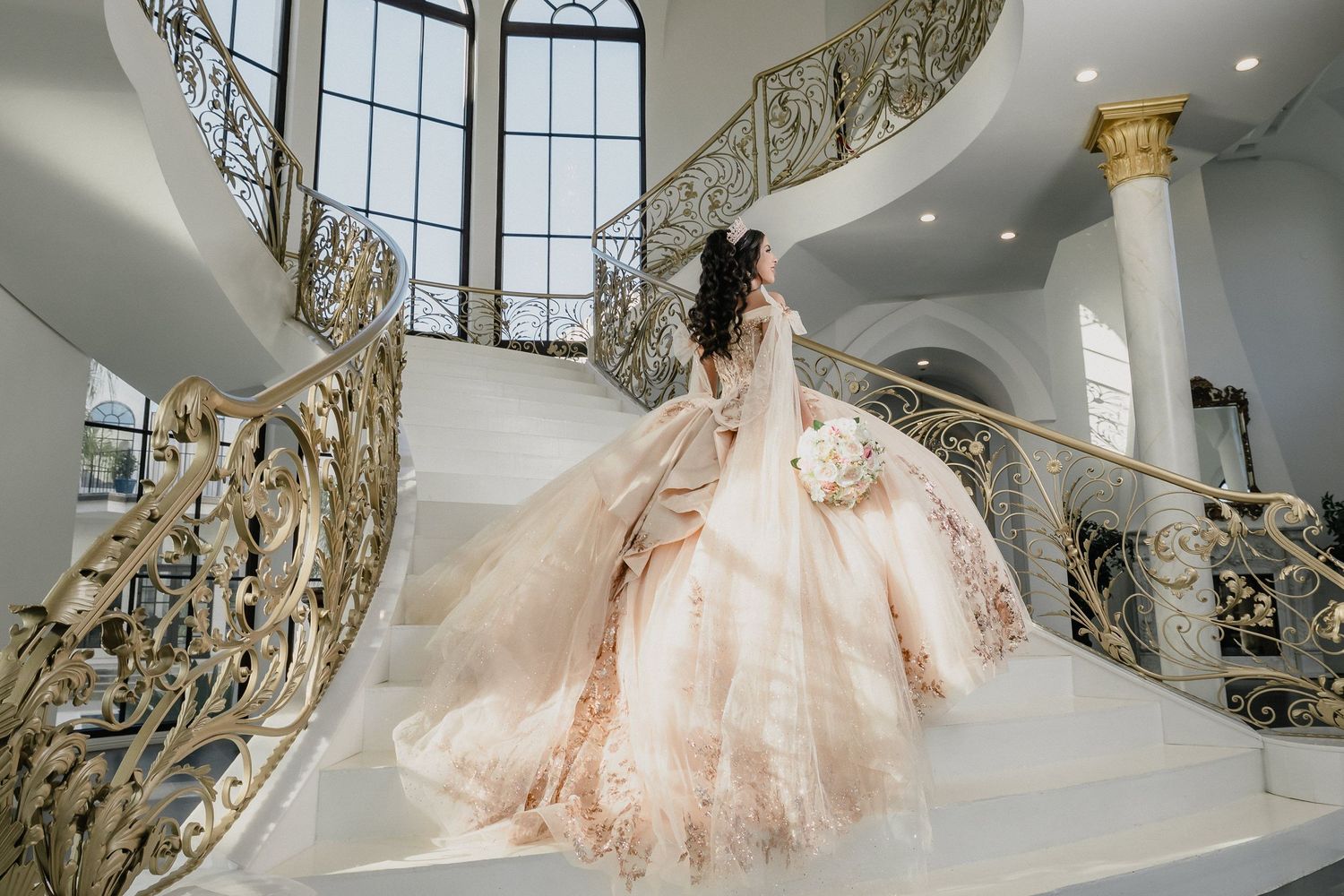 A flowing champagne ballgown captured on a grand white staircase with ornate gold railings.