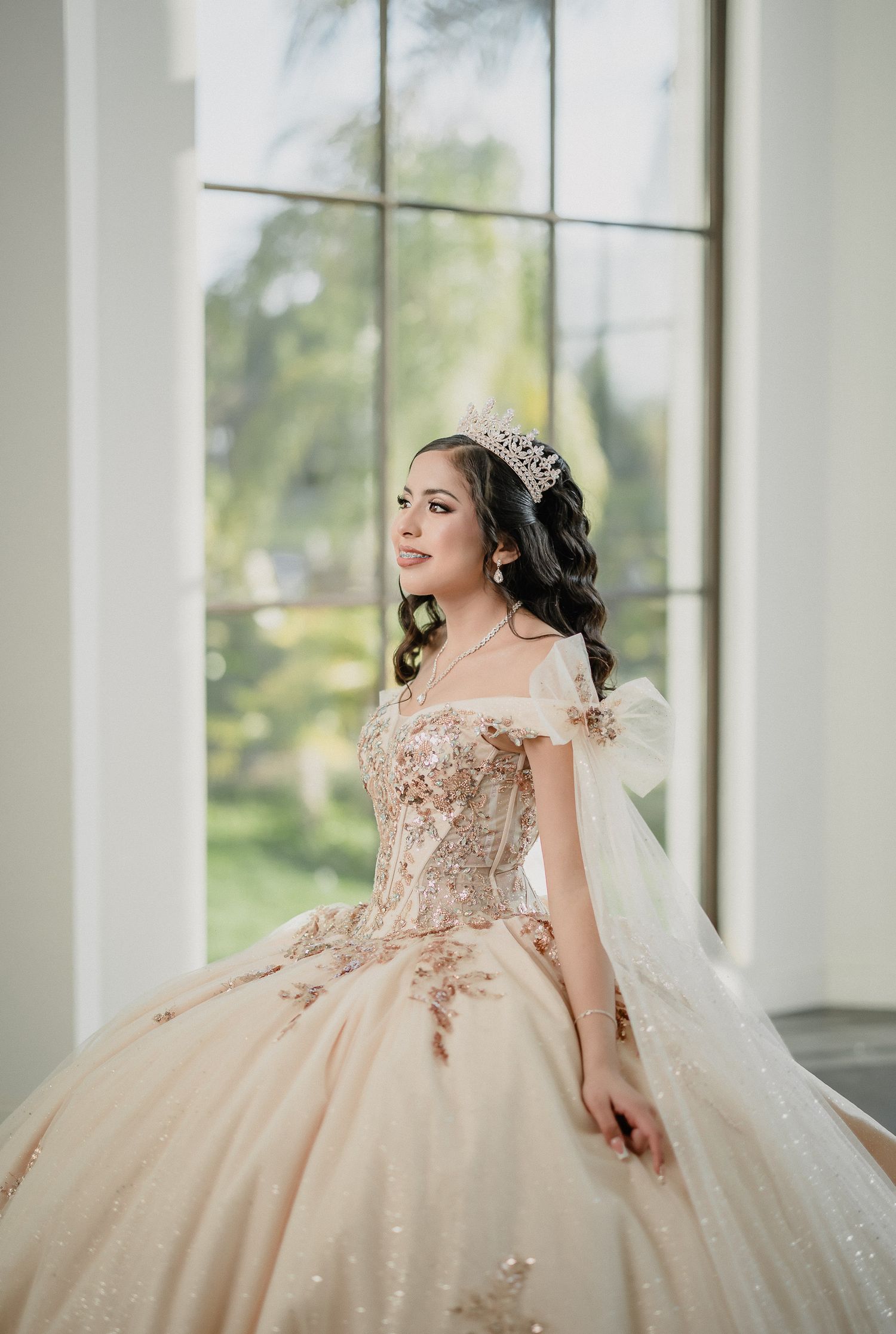 Beautiful quinceanera dress in champagne color with off-shoulder design and delicate floral posing by a window.