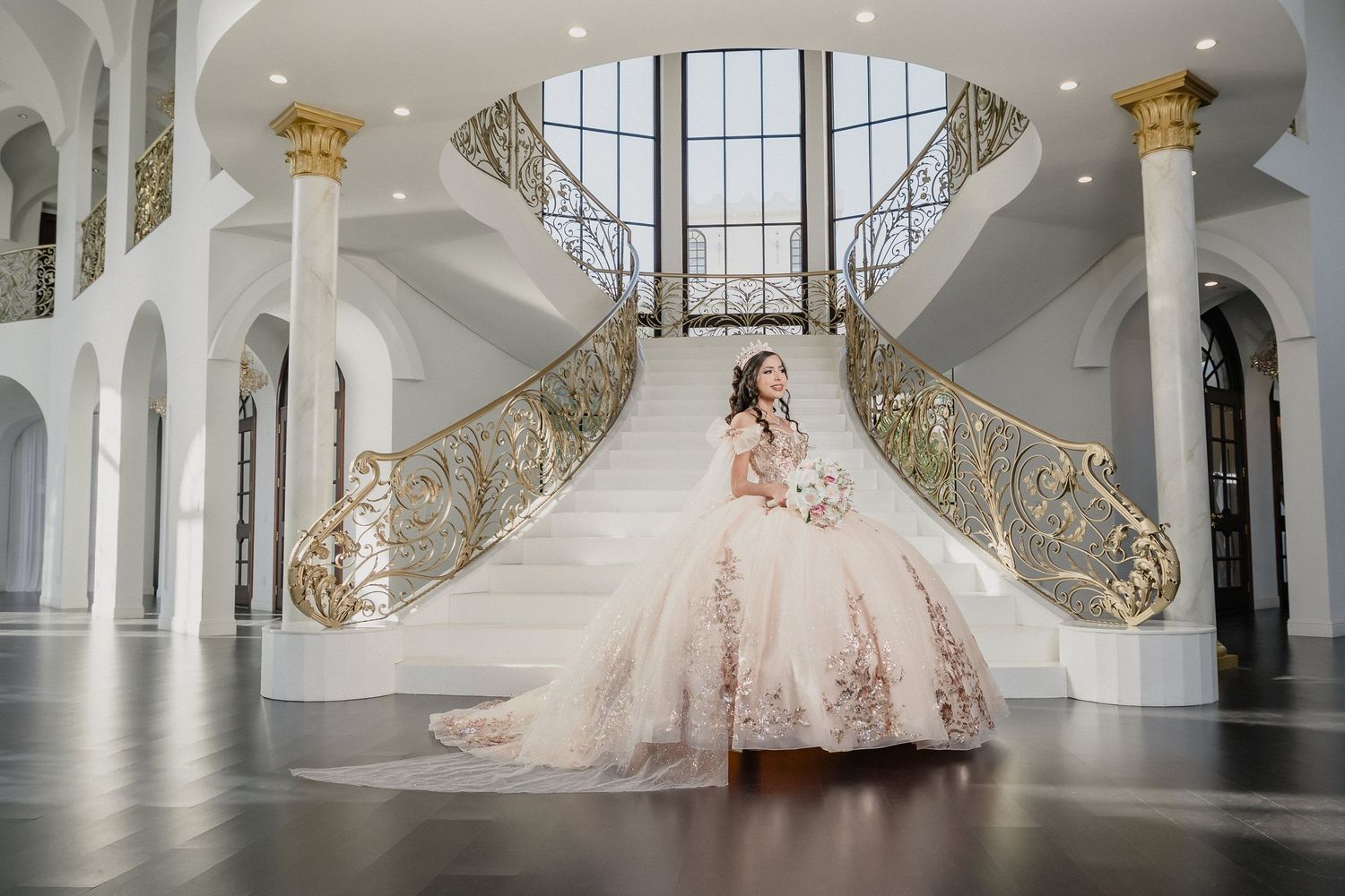 An elegant figure in a blush pink quinceanera poses before grand dual staircases with ornate gold railings in a luxurious venue.