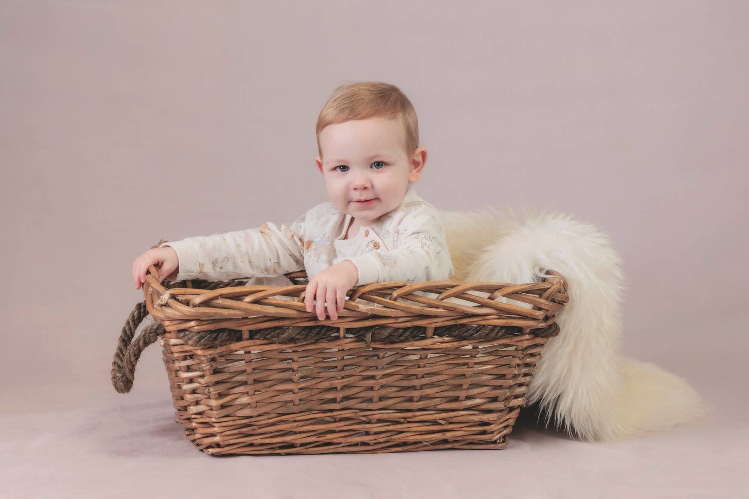A small child sits in a rustic wicker basket with a white furry blanket . Tiny moments photography, baby & newborn hampshire photographer