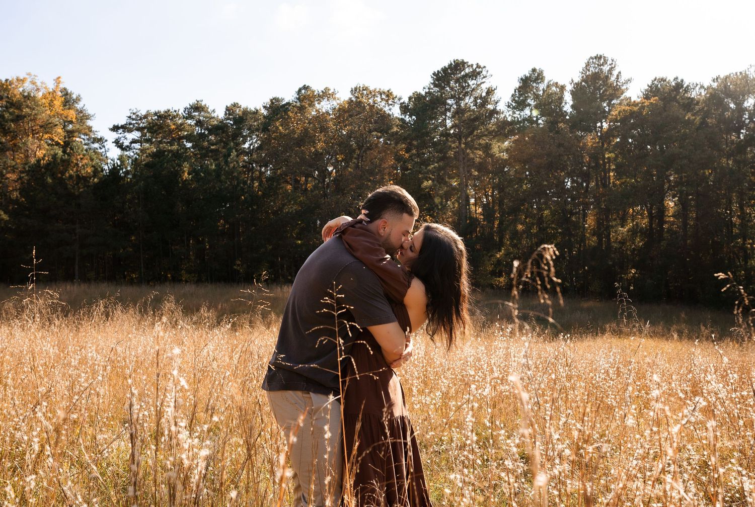 Romantic couple embracing in sunny autumn field with trees in background.