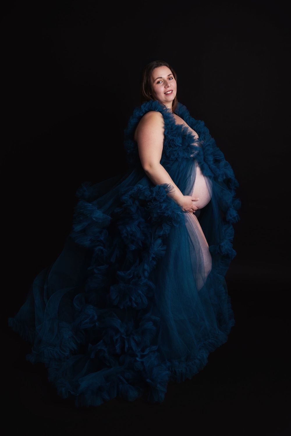 Dramatic maternity portrait featuring a flowing dark blue tulle gown against a black background.