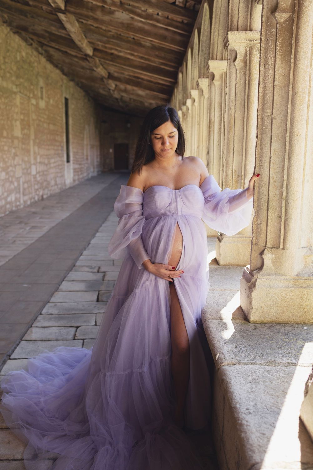 A pregnant woman in a flowing lavender gown poses in an elegant stone corridor with dramatic natural lighting.
