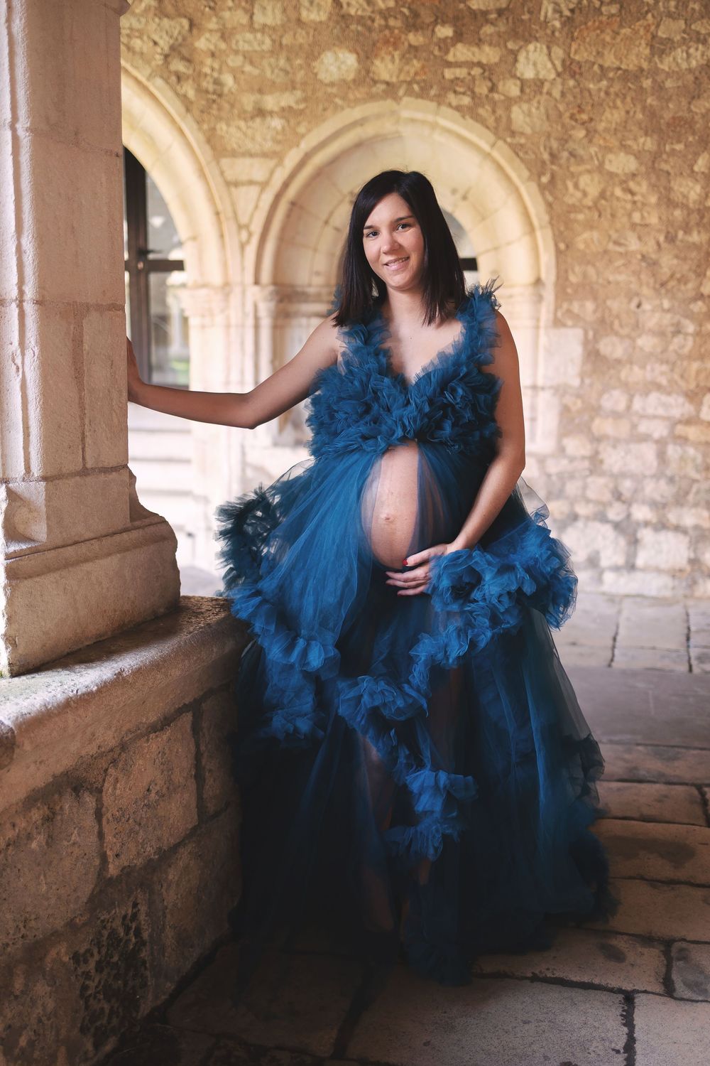 An expectant figure poses in a dramatic blue ruffled gown with tulle details against a stone archway backdrop.