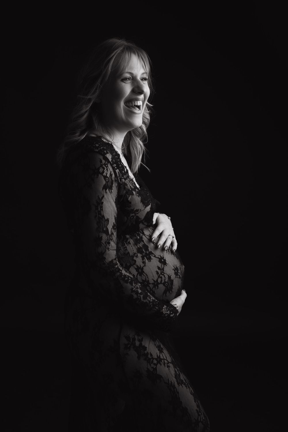 Black and white maternity portrait of an expectant mother in lace dress posing against dark background.