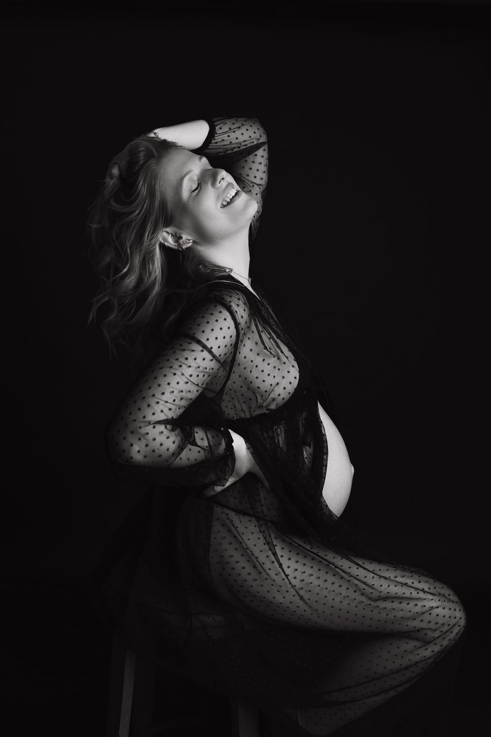 Artistic black and white portrait of a person in sheer polka dot bodysuit striking dramatic pose against dark backdrop.