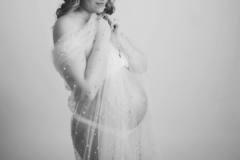 Black and white maternity portrait with delicate lace veil creating an ethereal silhouette against light background.
