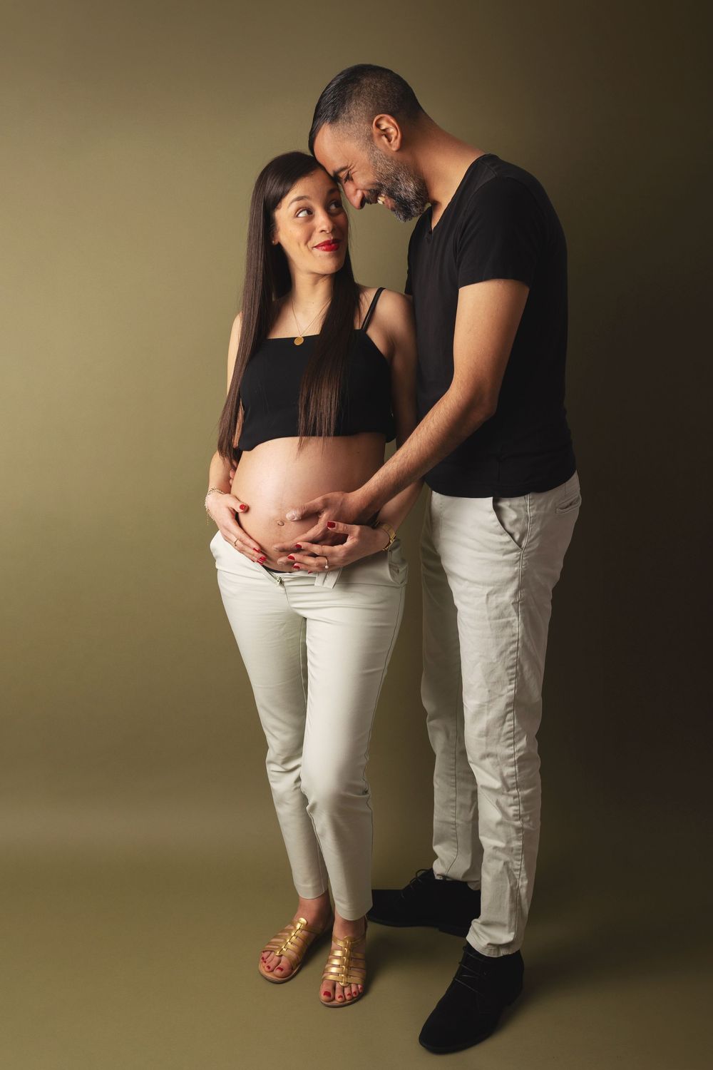 Expecting couple wearing white pants and black tops share tender moment during maternity photoshoot against beige backdrop.