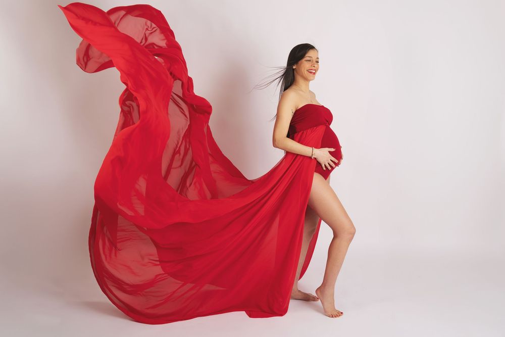Flowing red chiffon maternity gown billows dramatically against white backdrop in elegant pregnancy photo shoot.