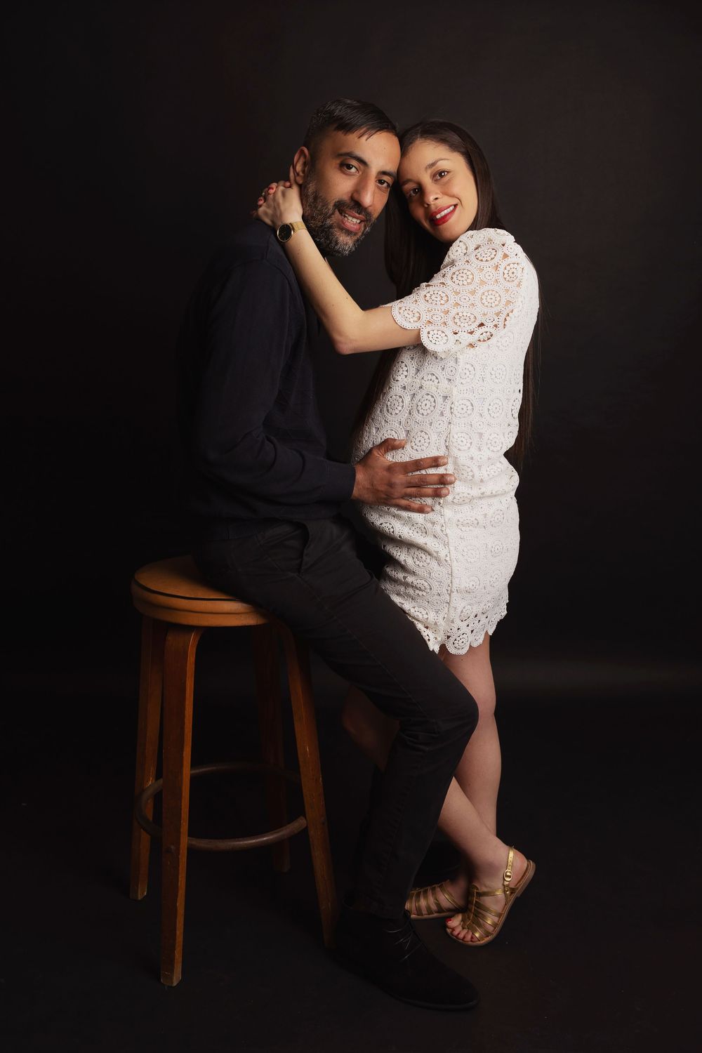 A romantic couple poses intimately against a dark background, with her wearing white lace and him seated on a wooden stool.