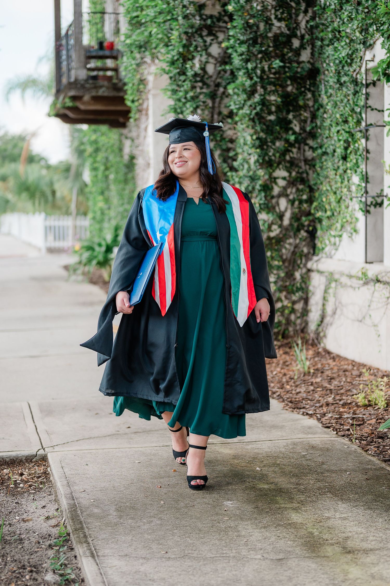 Graduate in emerald green dress and academic regalia walks along an ivy-covered pathway.