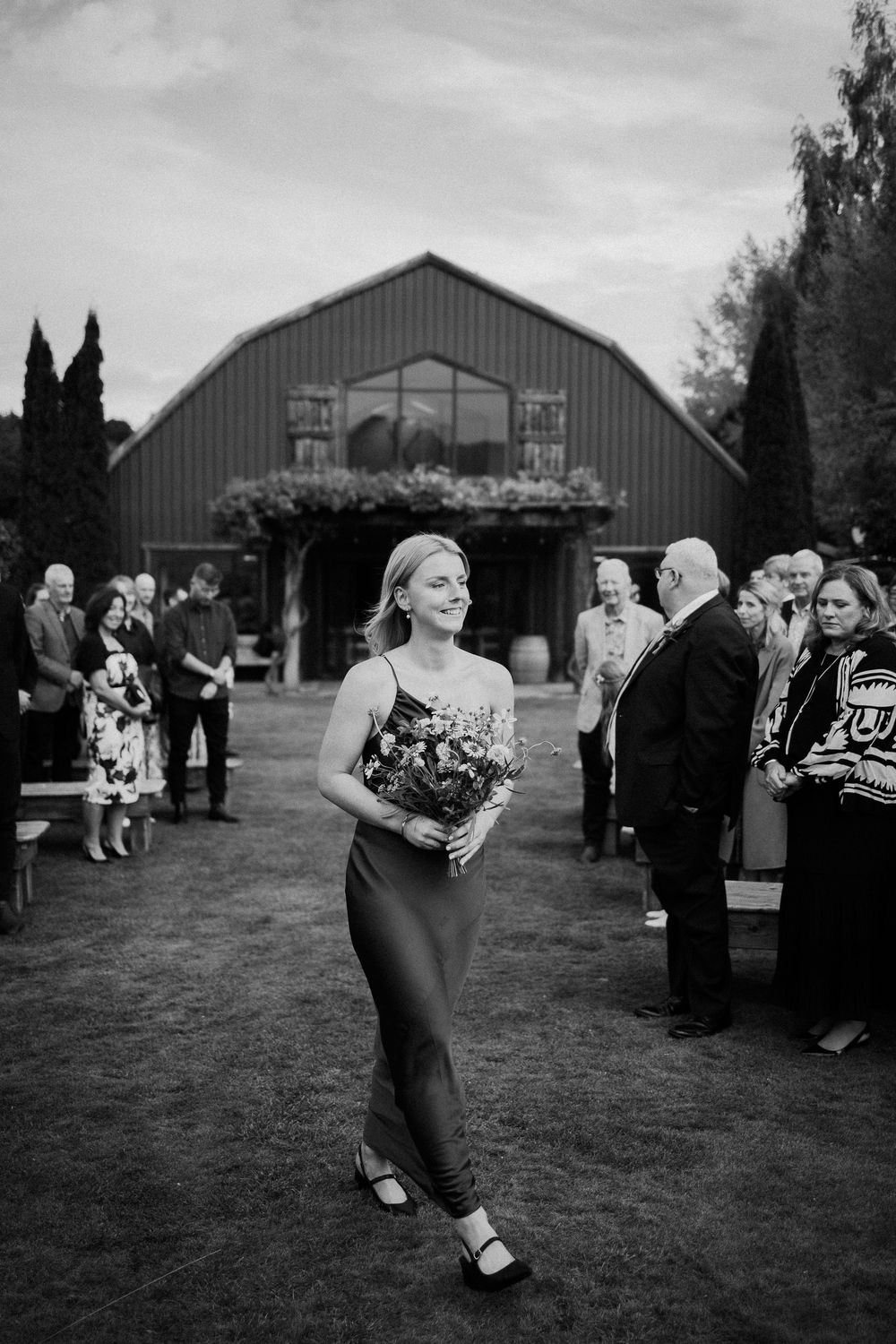 A black and white wedding photo shows guests gathered outside a rustic barn venue while someone walks down the aisle.