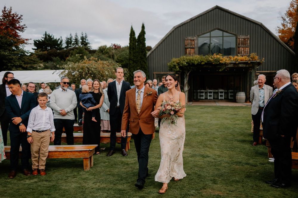 A rustic barn wedding ceremony outdoors with guests lined up on both sides as a couple walks down the grassy aisle.