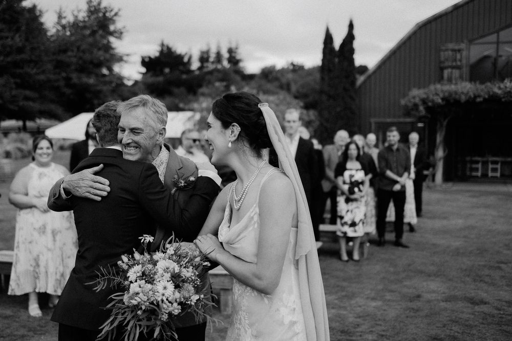Black and white photo of a tender wedding moment outside a rustic barn with guests gathered in the background.