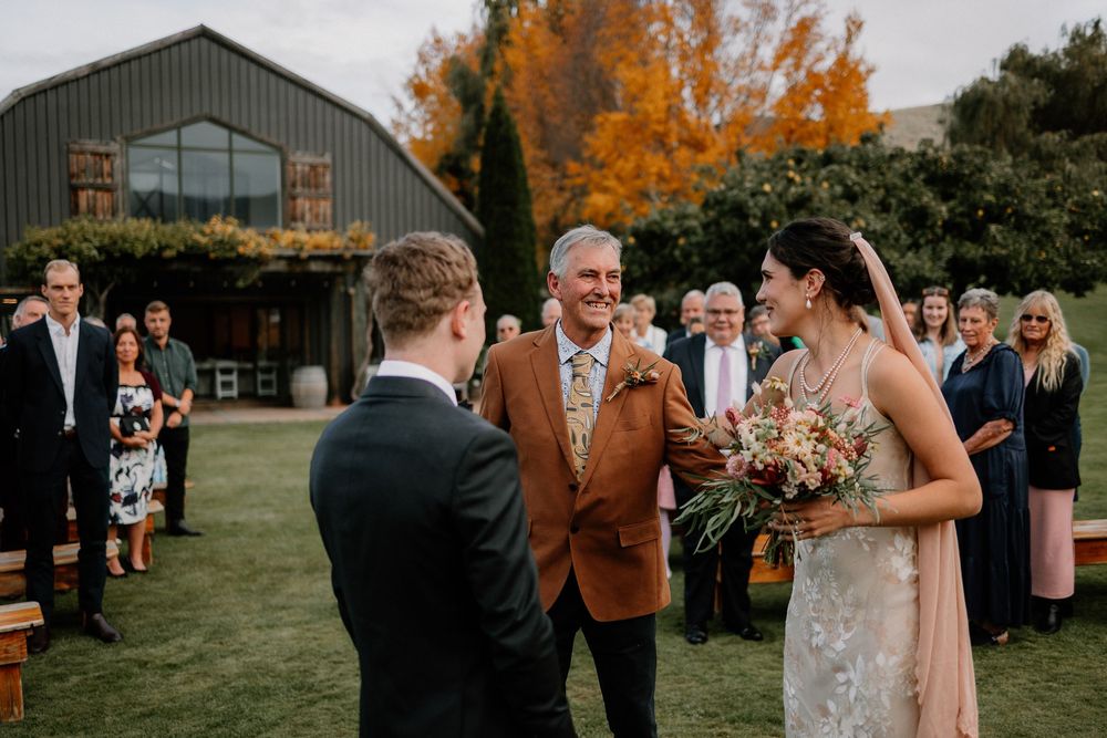 Outdoor wedding ceremony at a rustic barn venue with autumn trees and guests gathered on the lawn.