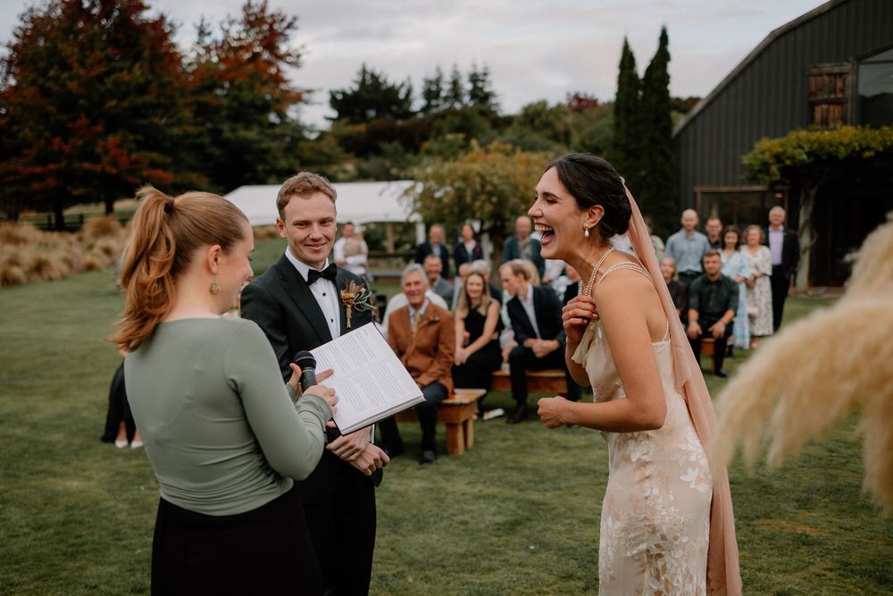 An outdoor wedding ceremony takes place on a lush green lawn with guests seated under autumn trees.