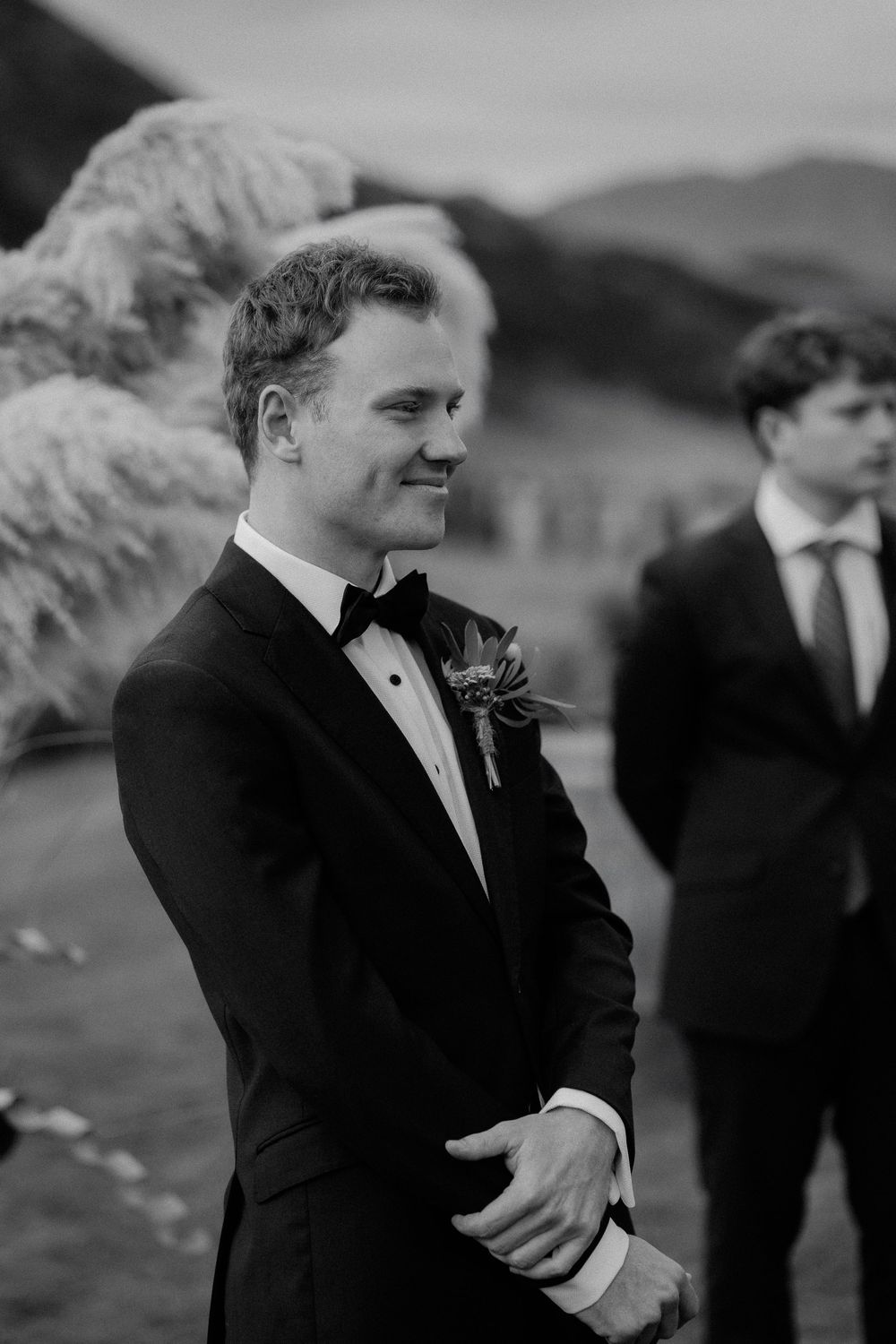 A groom in a classic black tuxedo with boutonniere smiles during an outdoor wedding ceremony.