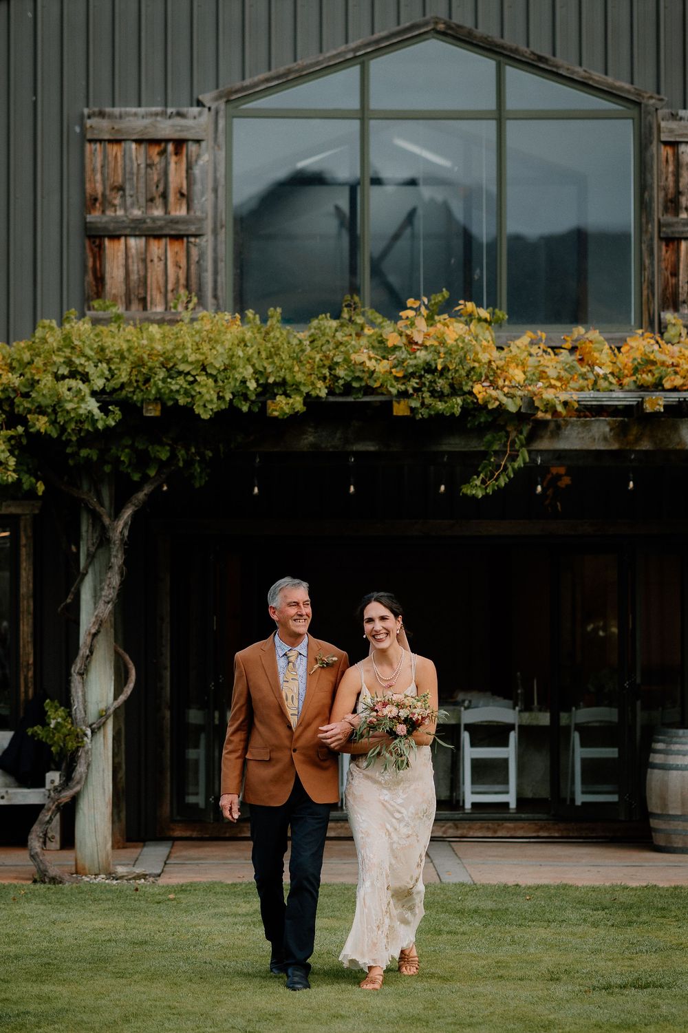 Two people walk together on a green lawn outside a rustic barn venue with climbing vines and glass windows.