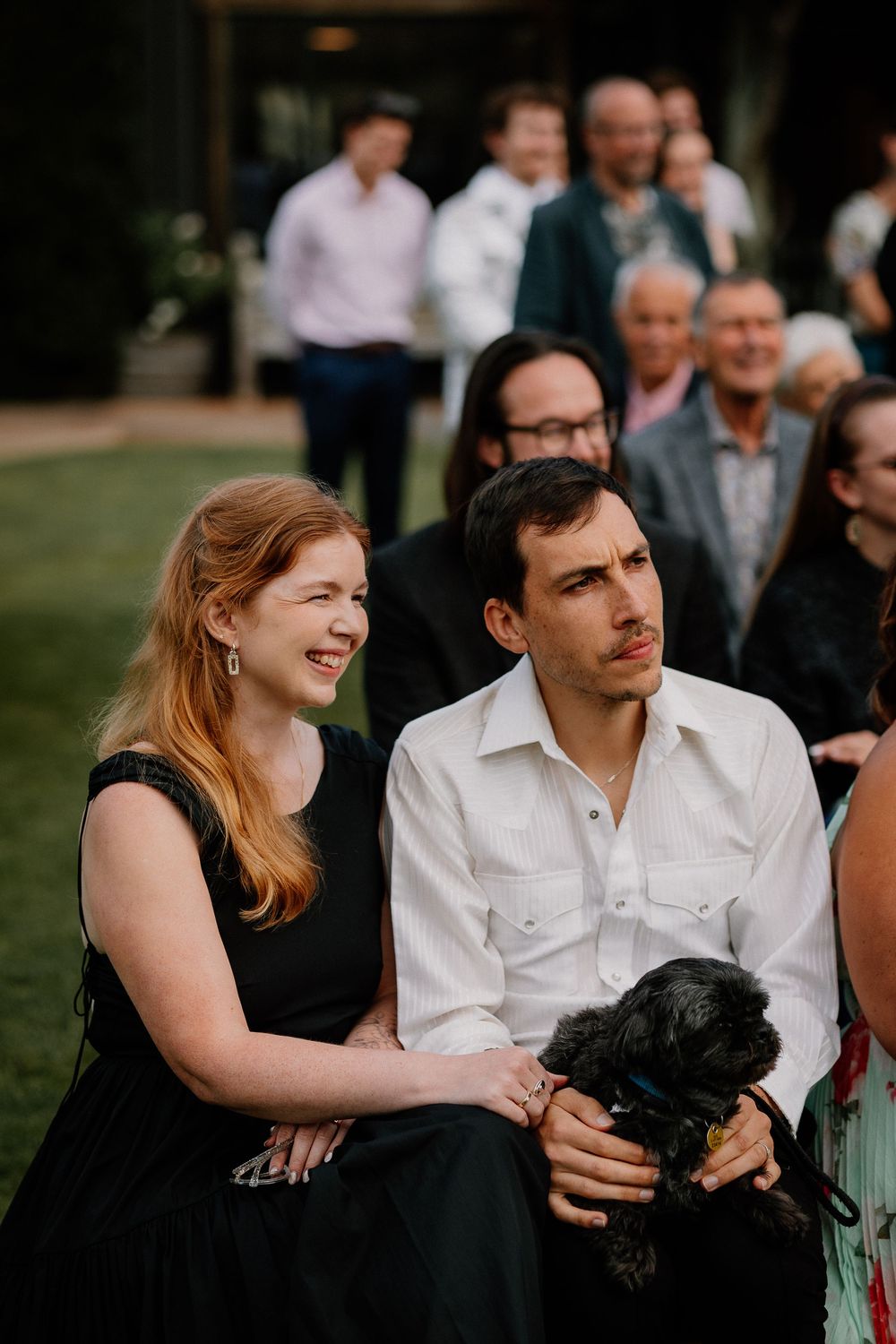 A couple sits outdoors at a gathering with a small black dog while others stand in the background.