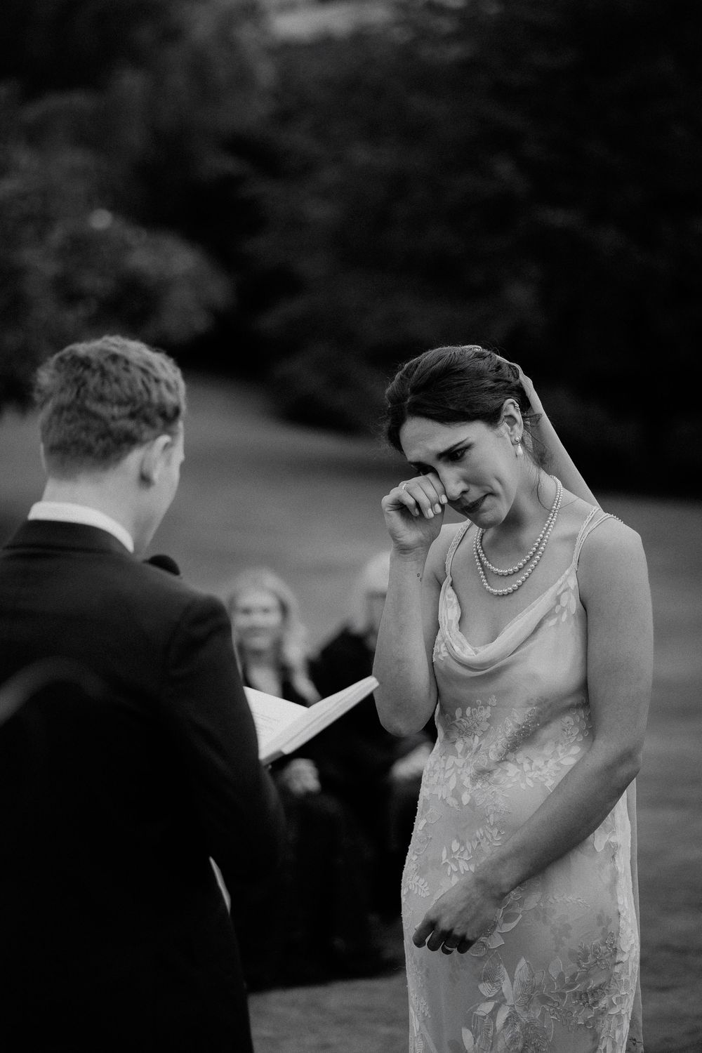 Emotional bride wipes away tears during a touching outdoor wedding ceremony as groom reads vows in black and white photography.