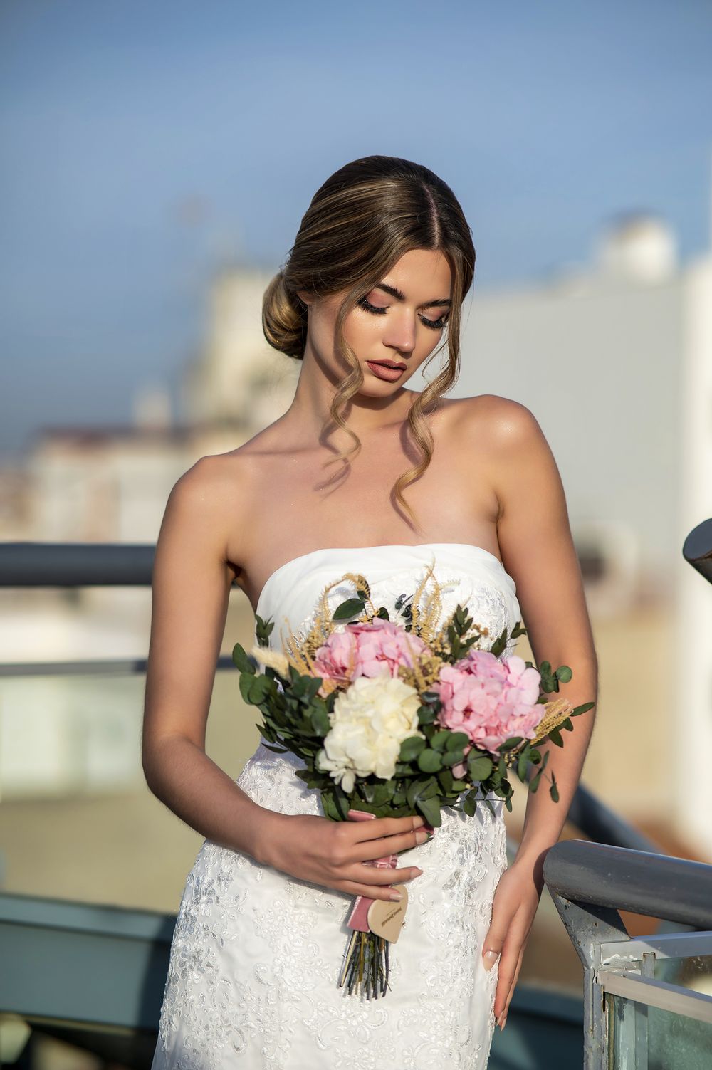 A bride in a strapless lace wedding dress holds a pink and white bouquet on a rooftop at sunset.