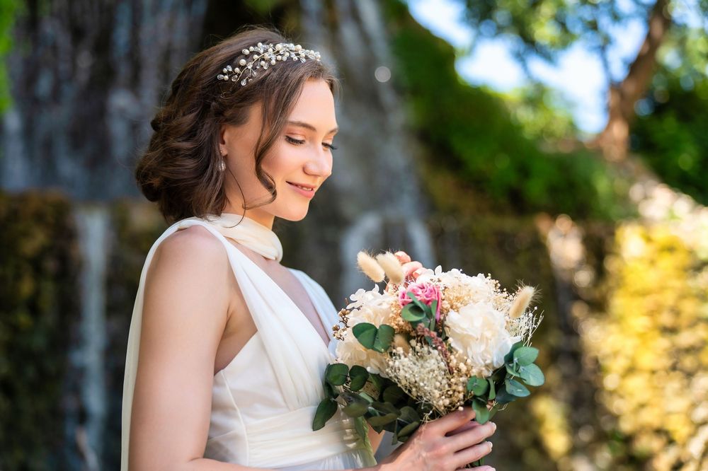 A bride in white dress with crystal hairpiece smiles while holding a pink and white floral bouquet in garden setting.