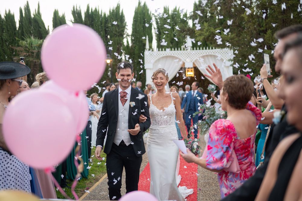 Wedding couple walks down aisle amid pink balloons and confetti while guests celebrate at outdoor garden ceremony.