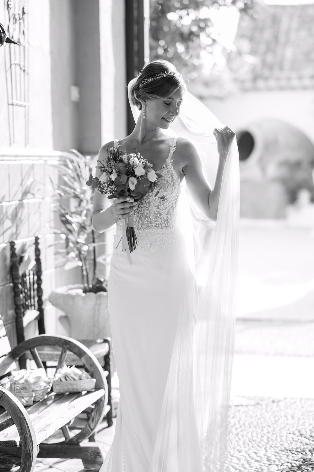 A bride in an elegant lace wedding gown holds her flowing veil while posing with a floral bouquet in black and white.