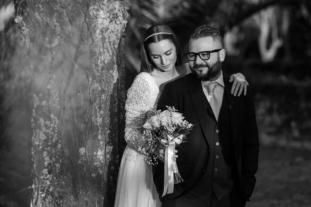 Black and white wedding photograph showing a romantic couple posing outdoors next to a tree.